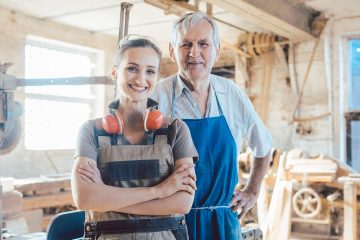 Senior master carpenter with his granddaughter in the wood workshop Senior master carpenter with his granddaughter in the wood workshop looking at camera