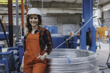 Portrait of young mechanical engineer woman working in factory building. Portrait of young happy apprentice machine operator woman checking component in a factory with white helmet smiling to camera and sitting in a large shipping manufacturing facility warehouse at metal cable factory XXXL